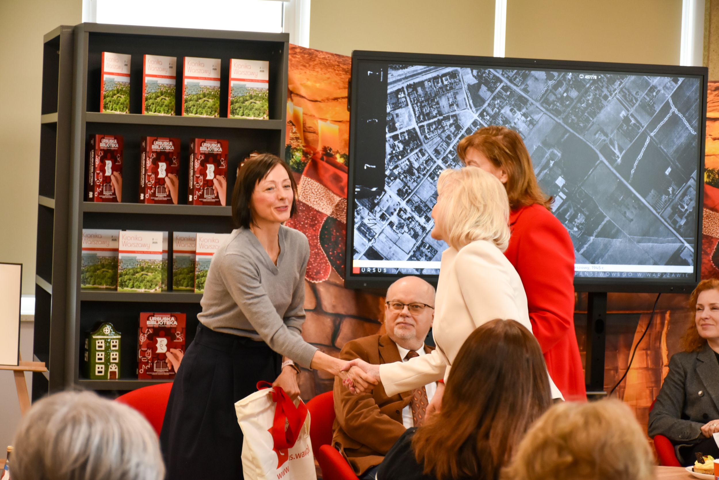 DSC_2010 Obok regału z książkami o Ursusie oraz przy tablicy dwie panie ściskają sobie dłonie. Kobieta na pierwszym planie trzyma w lewym ręku torbę z logo biblioteki. Reszta obserwujących uśmiecha się