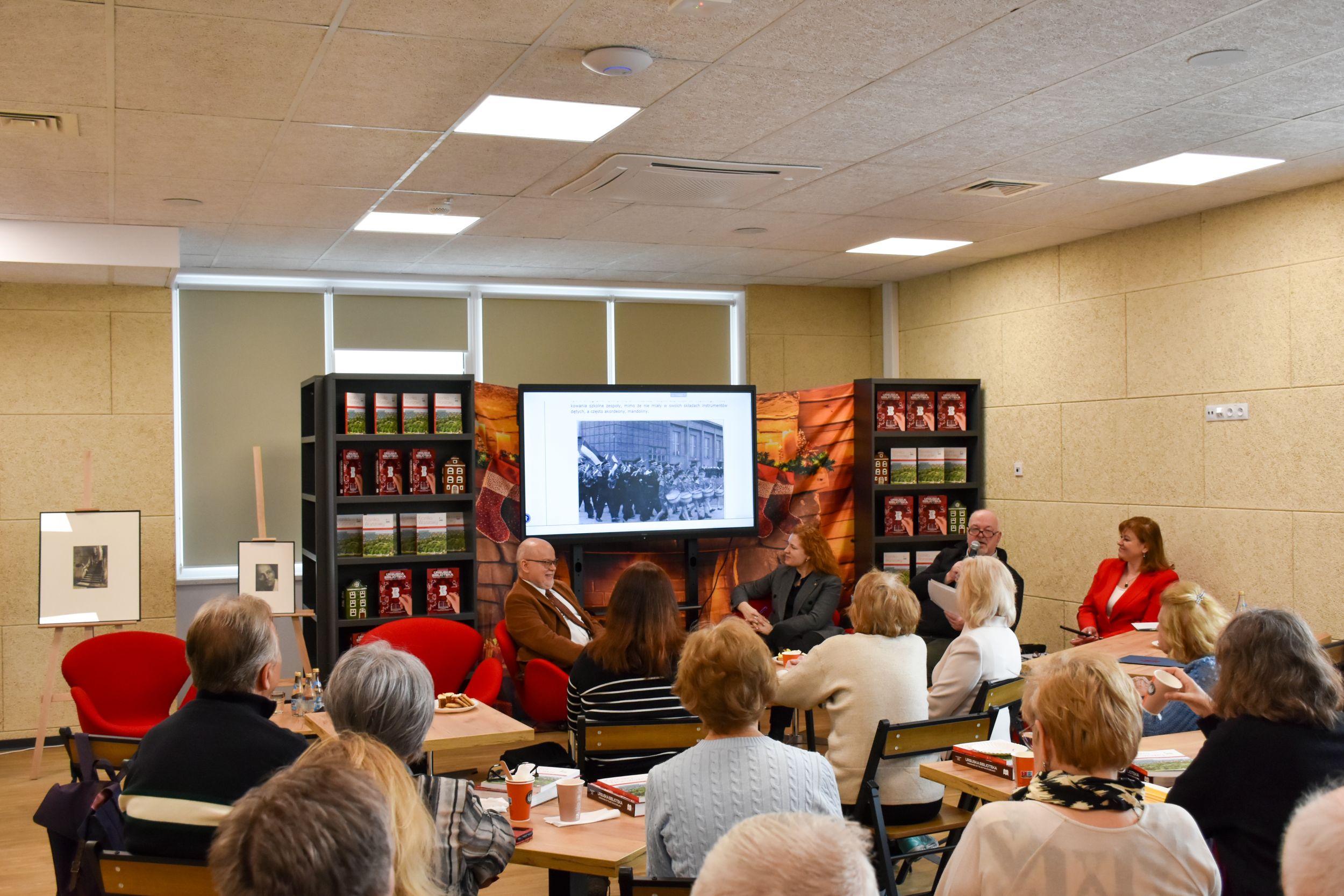 DSC_2019 Zdjęcie przedstawia grupę ludzi oglądających pokaz slajdów po obu stronach stoją regały z ursuskimi książkami