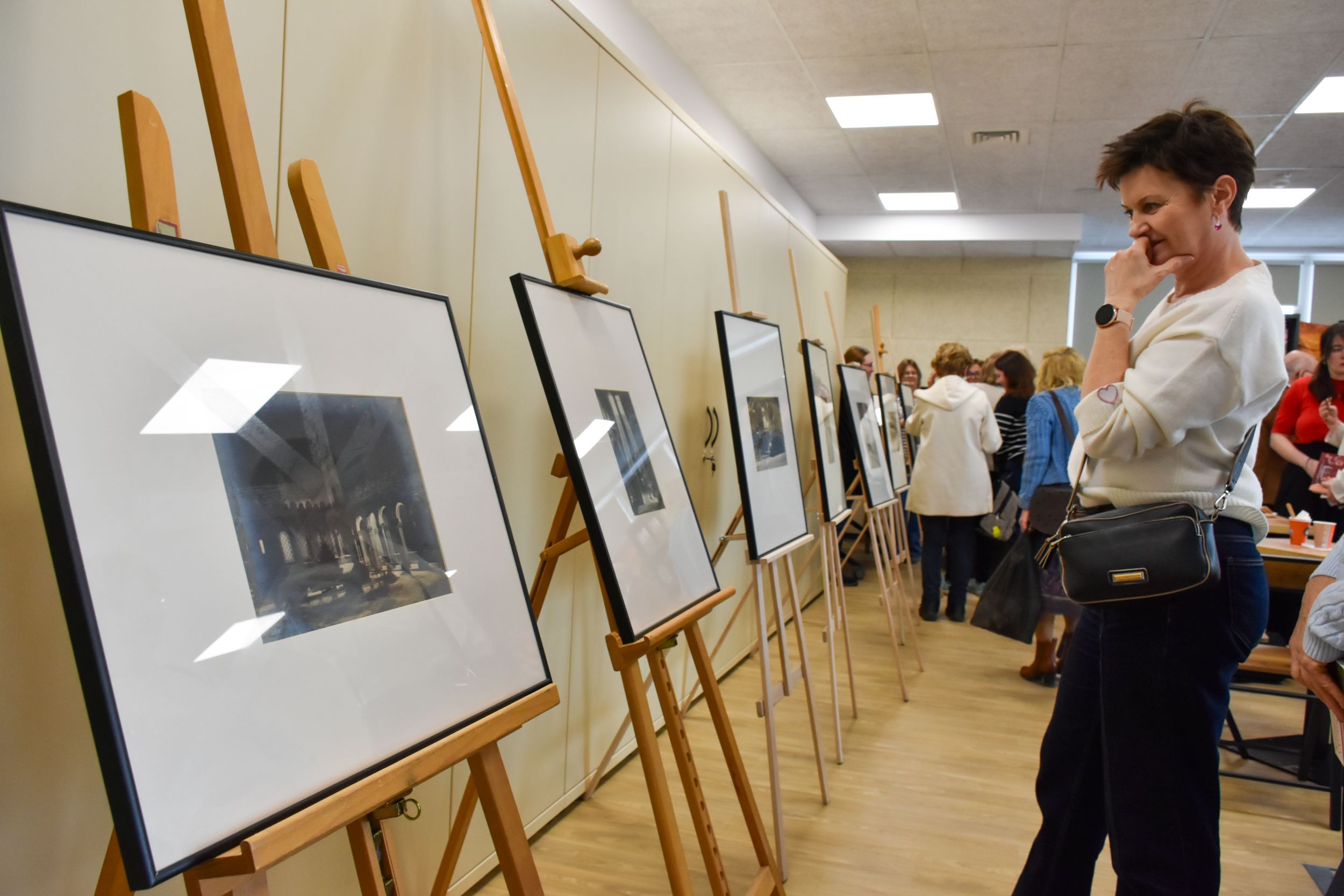 DSC_2078 Kobieta w białej bluzce i dżinsach z przewieszoną na ramię niewielką torebką z dłonią podpierającą podbródek ogląda z zaciekawieniem wystawę zdjęć. W rogu pomieszczenia stoi tłum rozmawiających ludzi