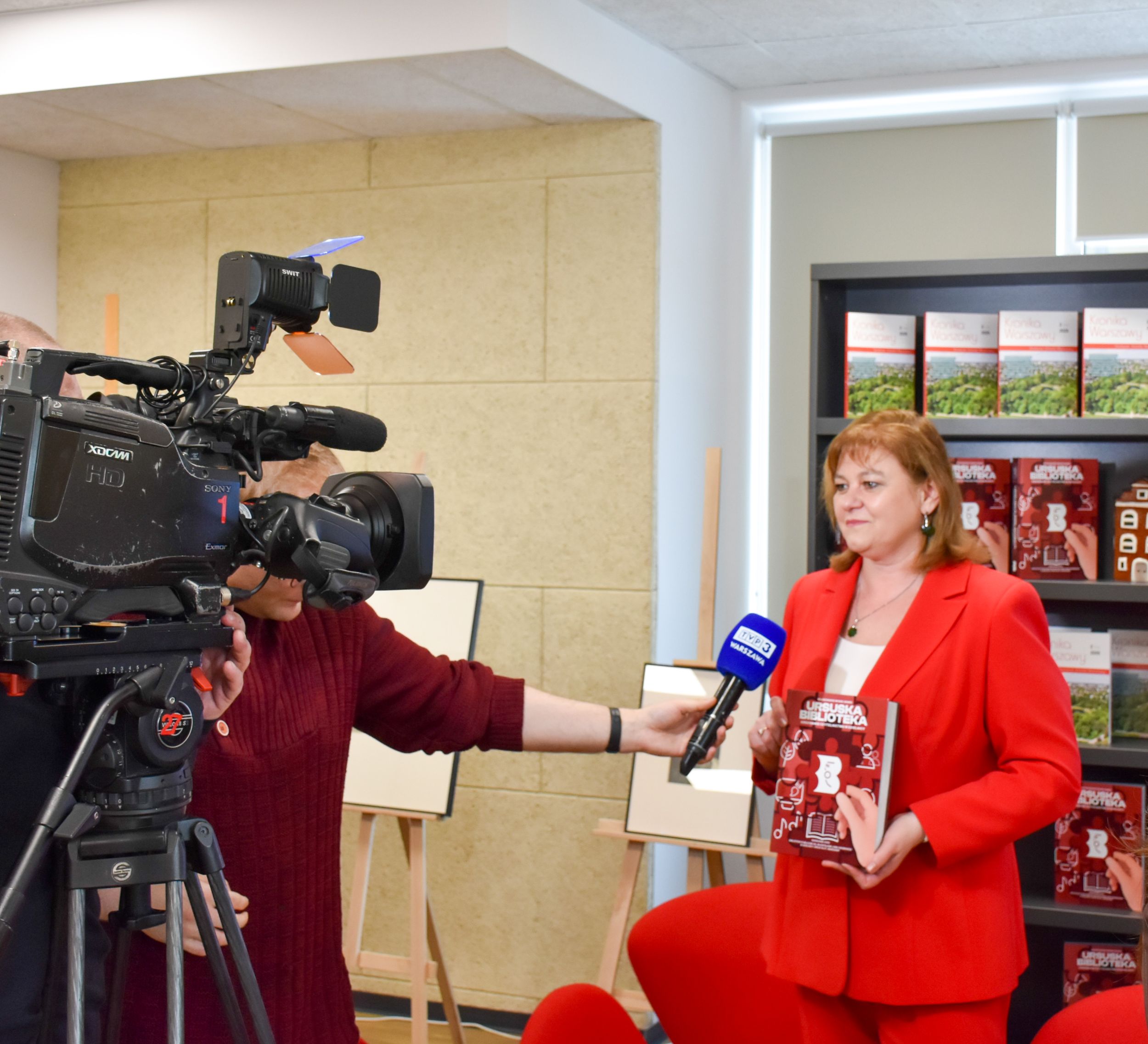 DSC_2088 Zdjęcie przedstawia kobietę w czerwonym kostiumie trzymającą książkę Ursuska Biblioteka która udziela wywiadu mężczyźnie z mikrofonem TVP 3 Warszawa po lewej stronie stoi zwrócona w stronę kobiety dużych rozmiarów kamera telewizyjna