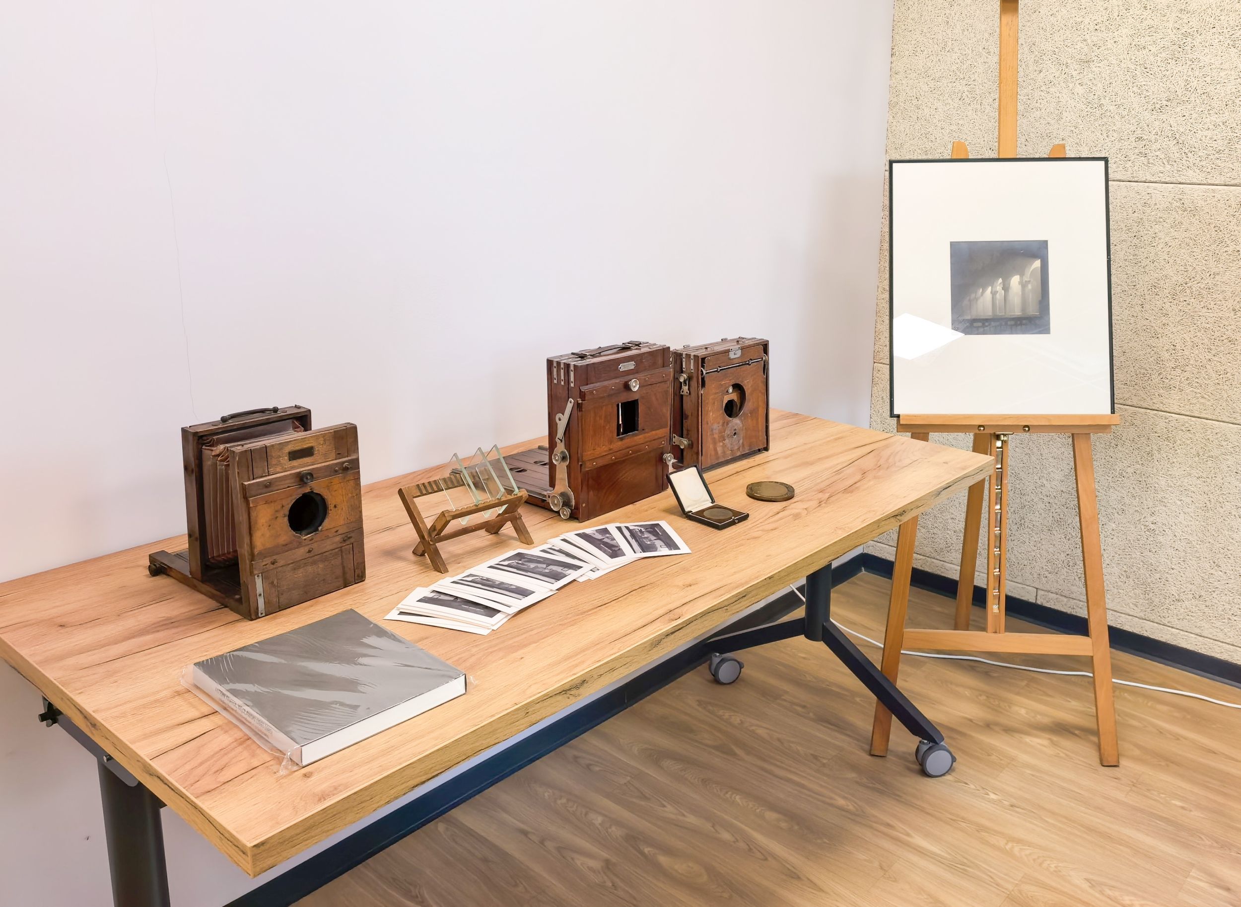 IMG_20260221_105717 Na stole stoją trzy kamery obscura pierwowzory aparatu fotograficznego wykorzystujące zjawisko optyczne polegające na rzutowaniu odwróconego obrazu rzeczywistości przez mały otwór na przeciwległą ścianę w ciemnym pomieszczeniu lub pudełku. Obok leżą pocztówki książka i medale oraz szkiełka