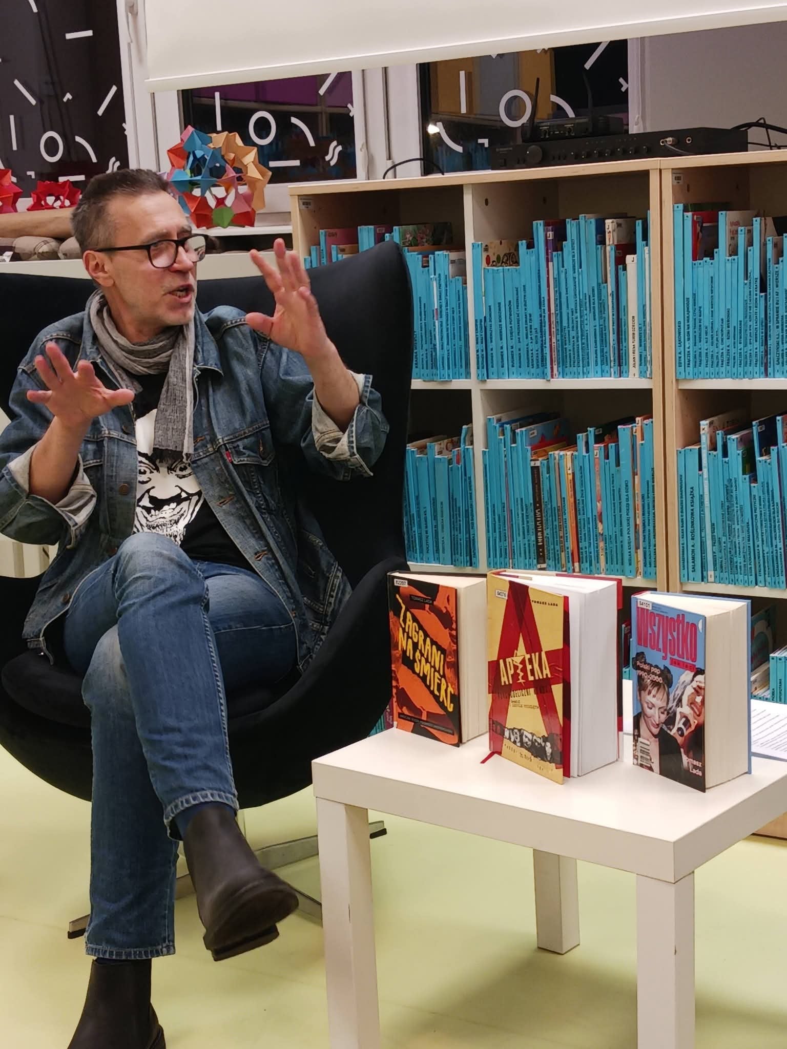 9bc9cc7c-722f-4220-85d1-d2d668561968 Mężczyzna w okularach siedzi na krześle, obok niego stół z książkami.