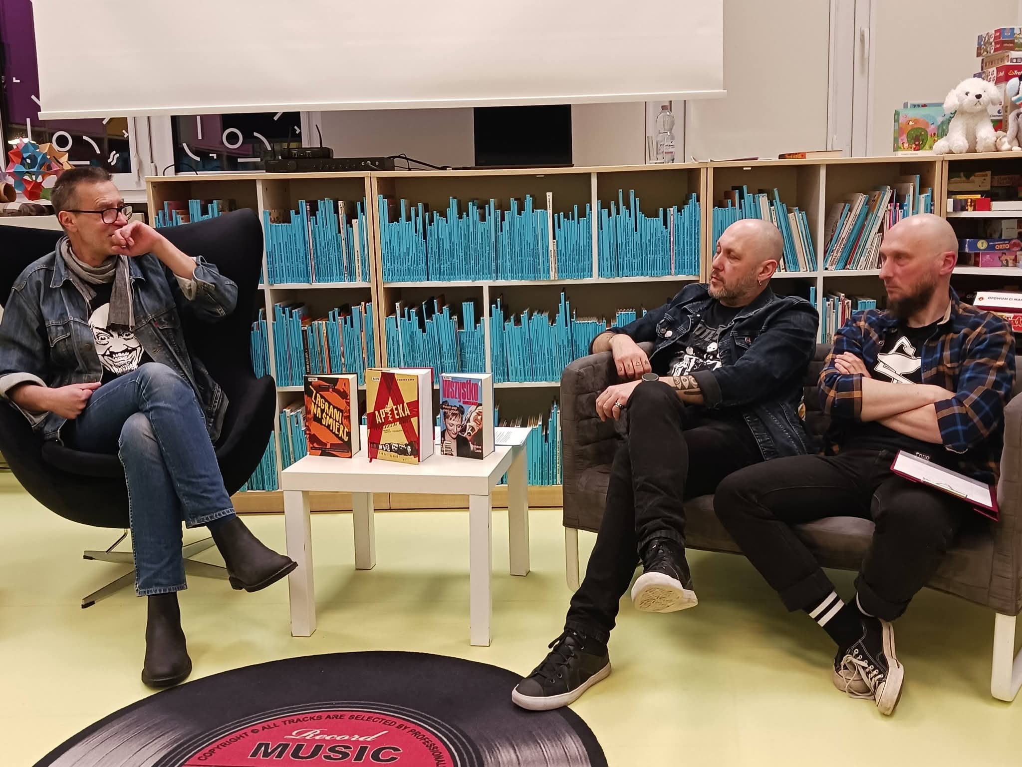 be6e7bb7-e358-4aab-9603-49f158fde392 Trzech mężczyzn siedzi na krzesłach przed regałem z książkami.