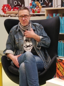 Mężczyzna w okularach siedzi na krześle, obok niego stół z książkami.