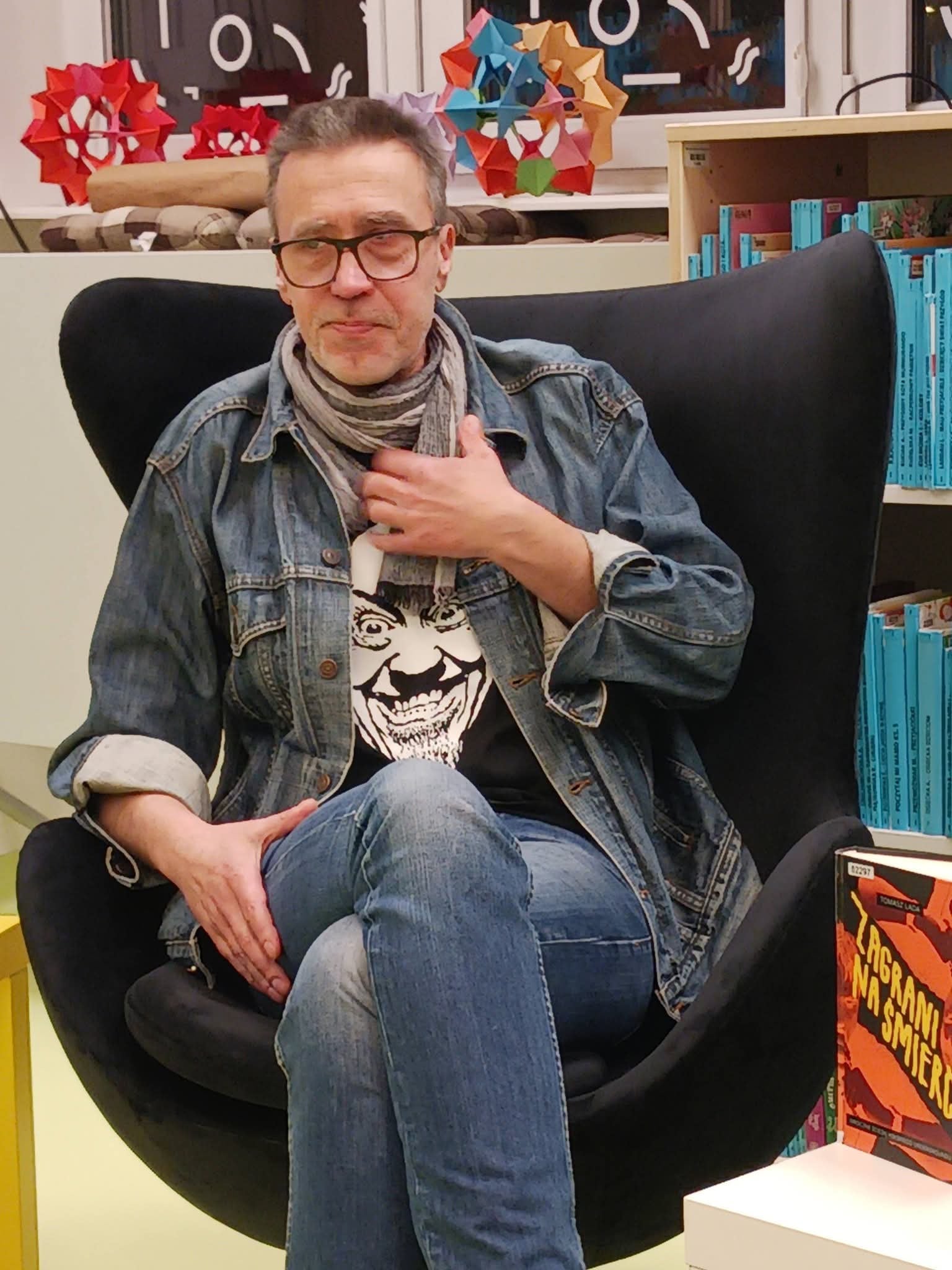 ca9b0d4e-d9fa-4114-978b-d21eb3722125 Mężczyzna w okularach siedzi na krześle, obok niego stół z książkami.