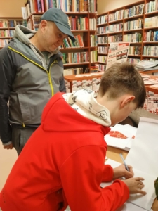 Mężczyzna i chłopiec przy, stojący przy stole. Chłopiec zapisuje coś na kartce.