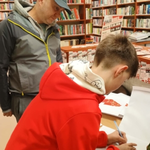 Mężczyzna i chłopiec przy, stojący przy stole. Chłopiec zapisuje coś na kartce.