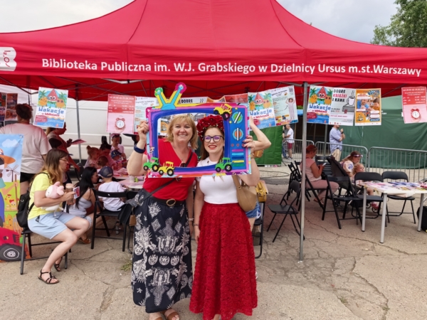 Dwie kobiety pozują do zdjęcia przed czerwonym namiotem, trzymając ramkę telewizora