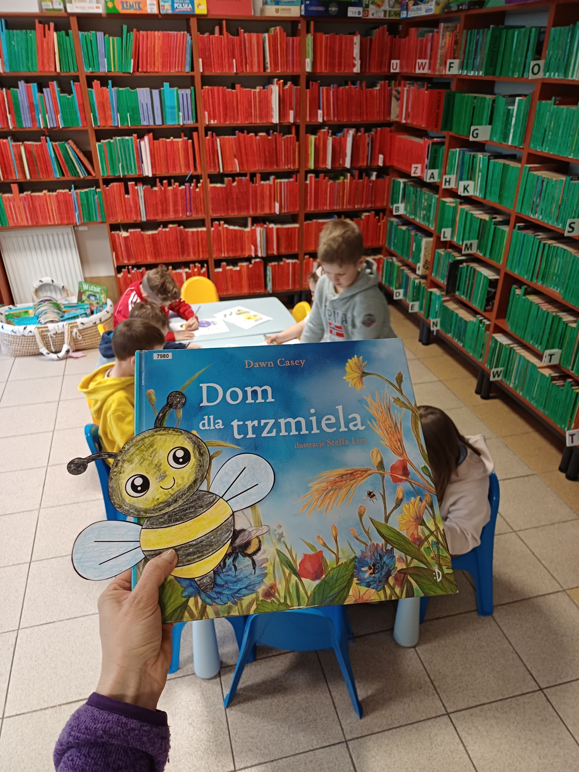 20260410_103435 Ręka z książką dziecięcą oraz dzieci, które siedzą przy stoliku.