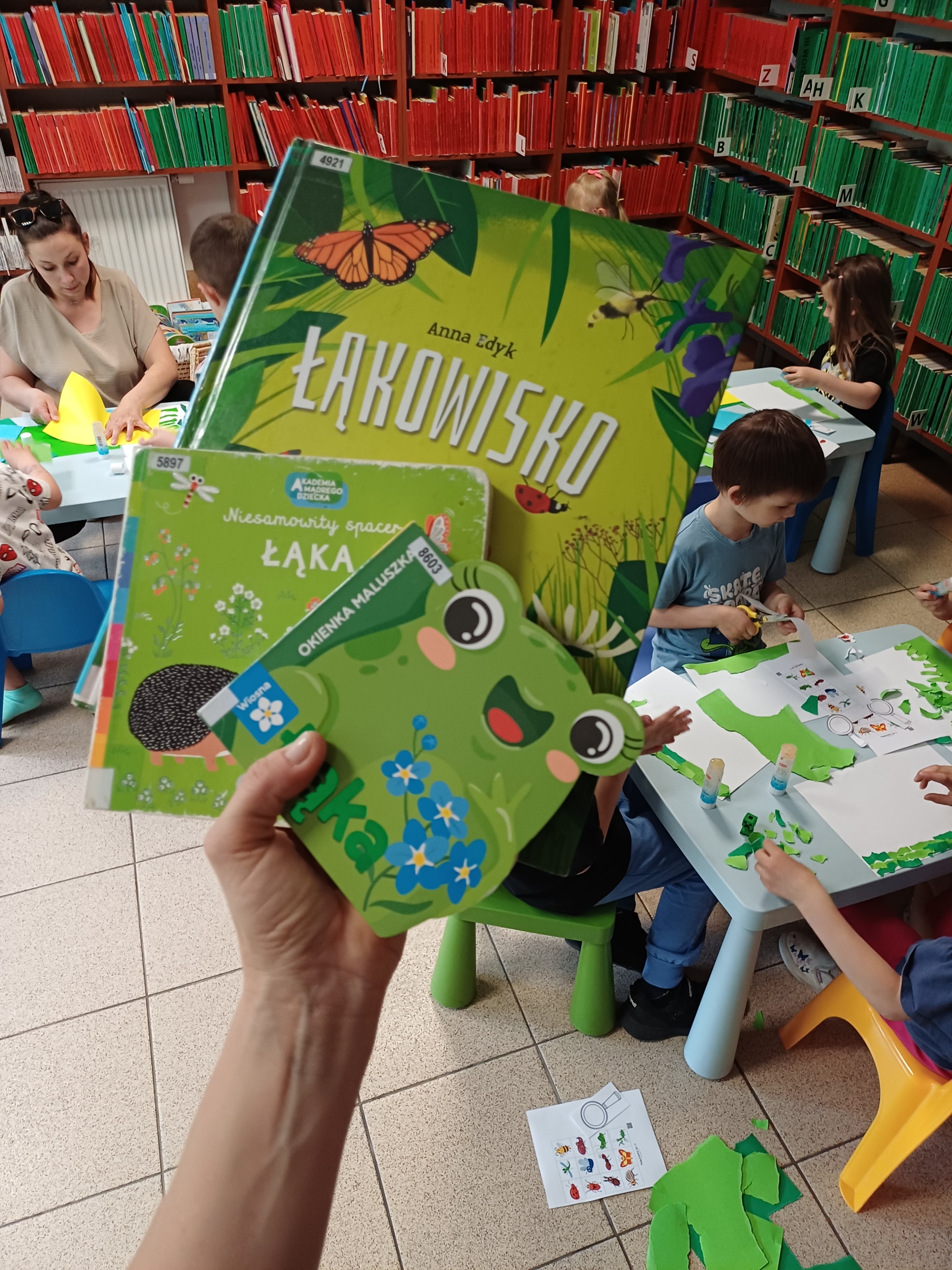 20260506_164005 Książki dla dzieci oraz dzieci i kobieta siedzący przy stolikach.