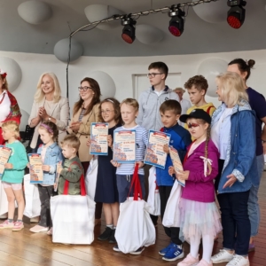 Dzieci i dorośli uśmiechają się na scenie, trzymając nagrody od biblioteki, pozując do wspólnego zdjęcia.