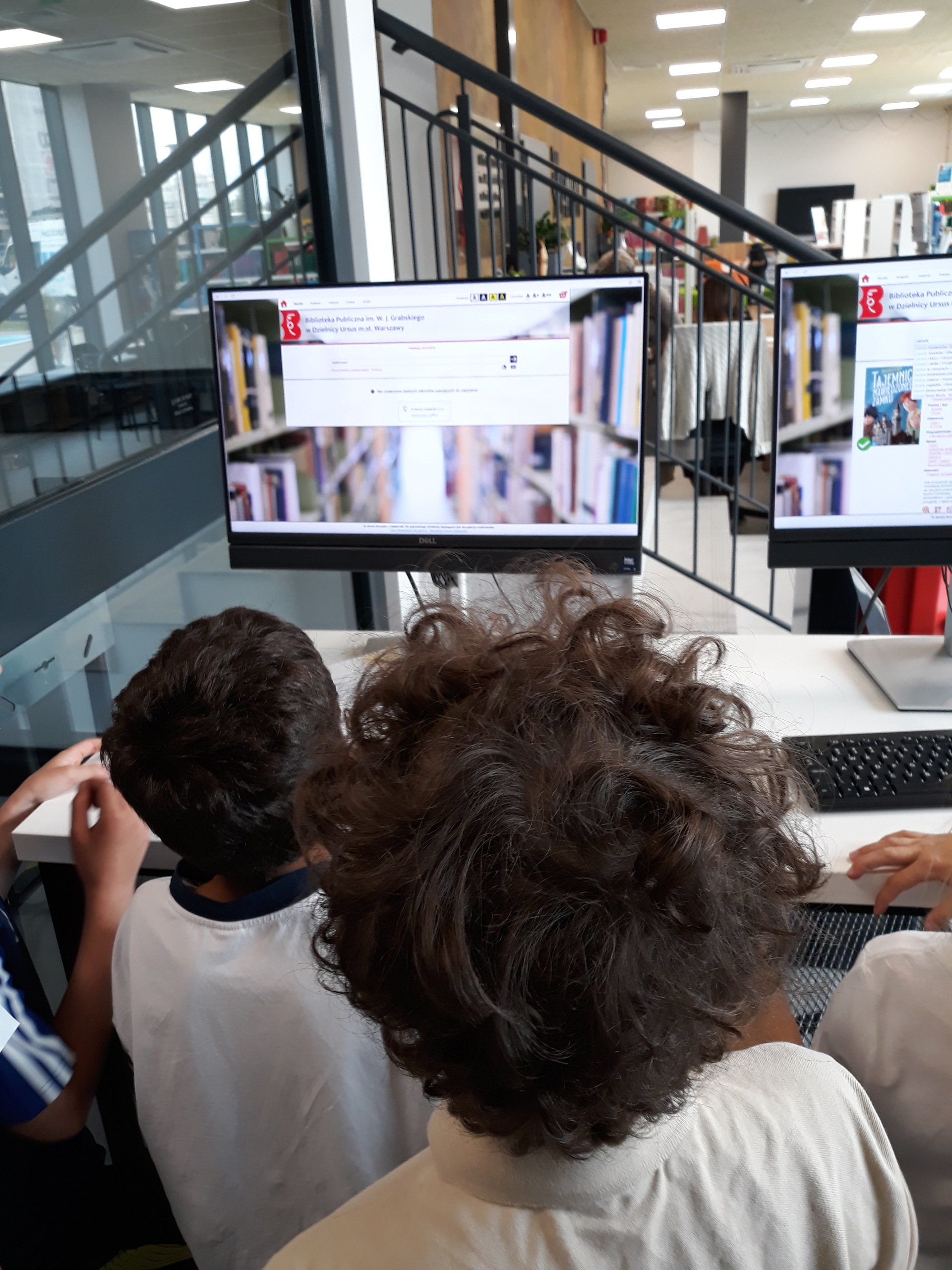 20260817_122016 Dzieci, przyglądające się wyświetlonym na monitorach stronom katalogu bibliotecznego.