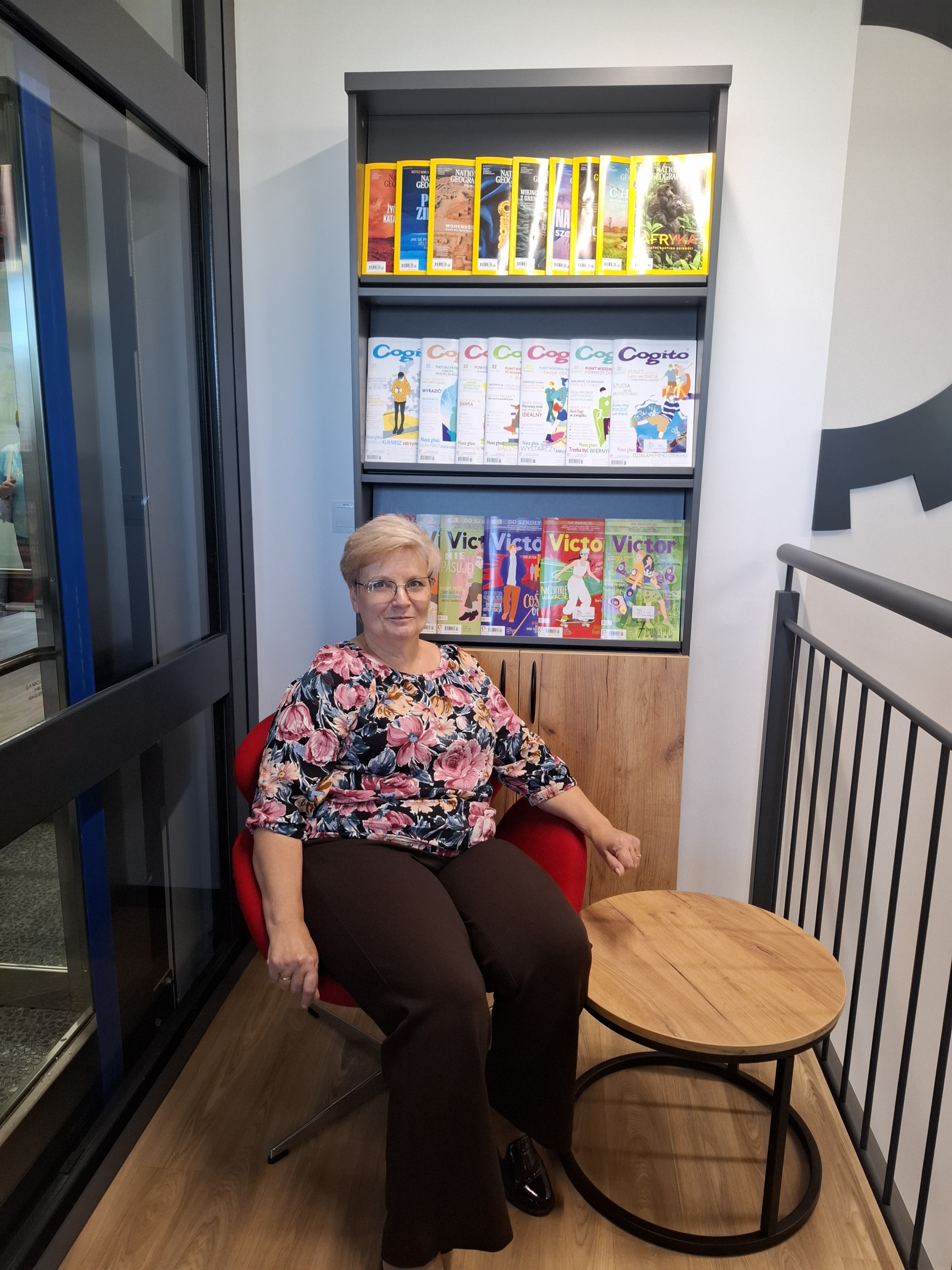 20260928_141929 Kobieta siedząca na krześle przed regałem z książkami, otoczona literaturą.