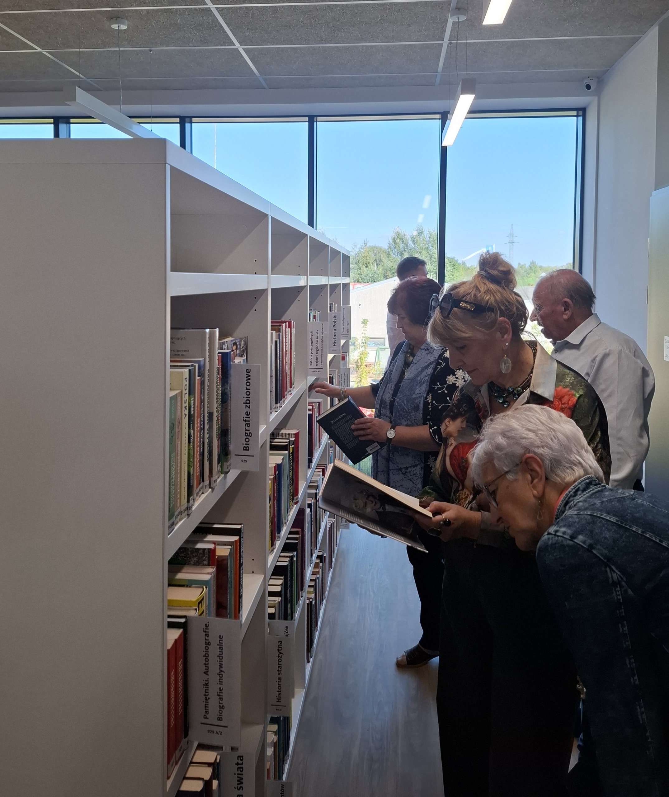 20260928_142309 Ludzie przeglądający książki w bibliotece, otoczeni regałami pełnymi literatury.
