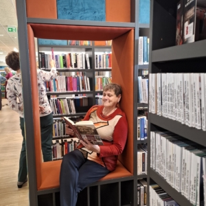 Kobieta siedząca w bibliotece obok regału z książkami, otoczona literaturą.