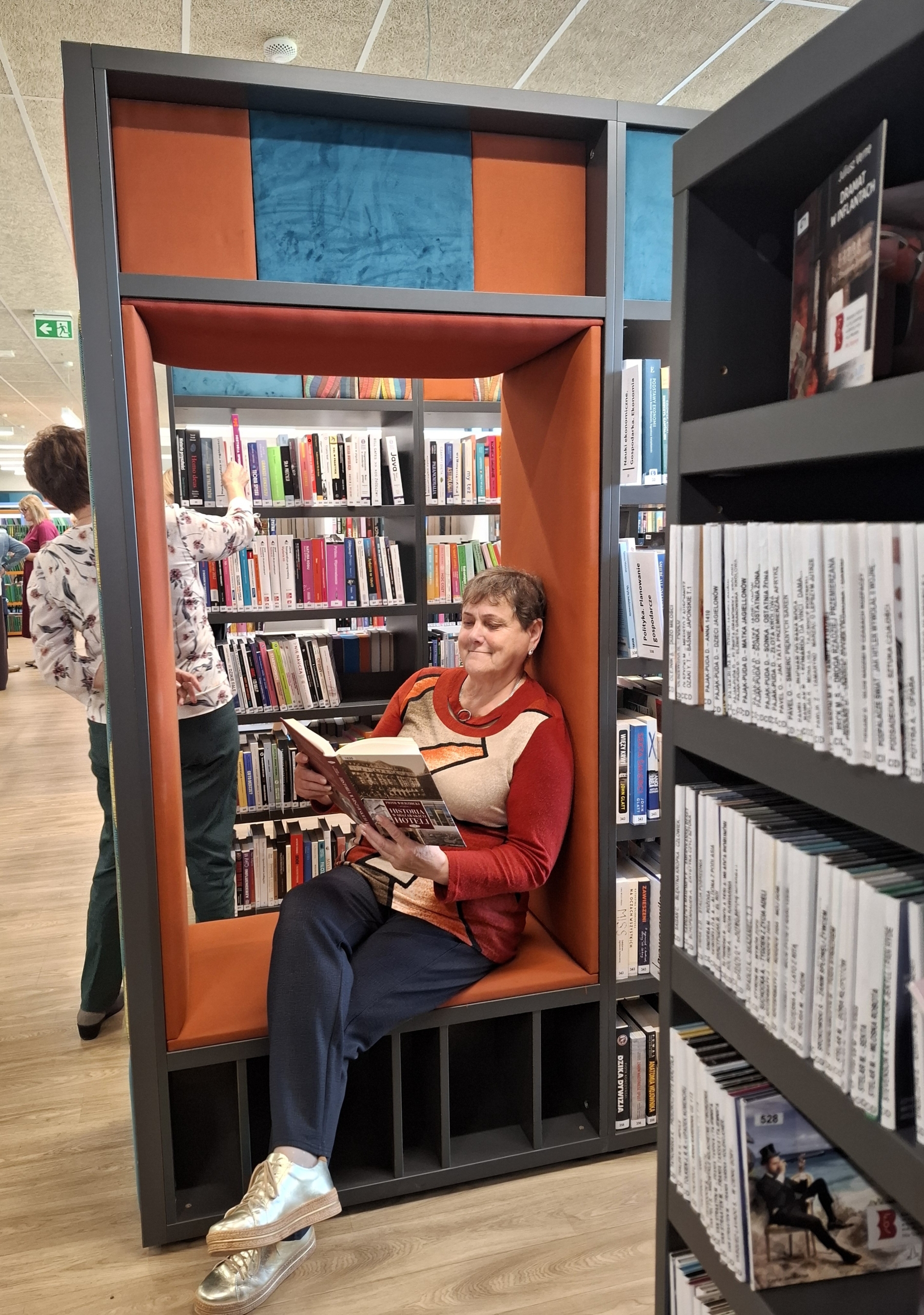 20260928_142329 Kobieta siedząca w bibliotece obok regału z książkami, otoczona literaturą.