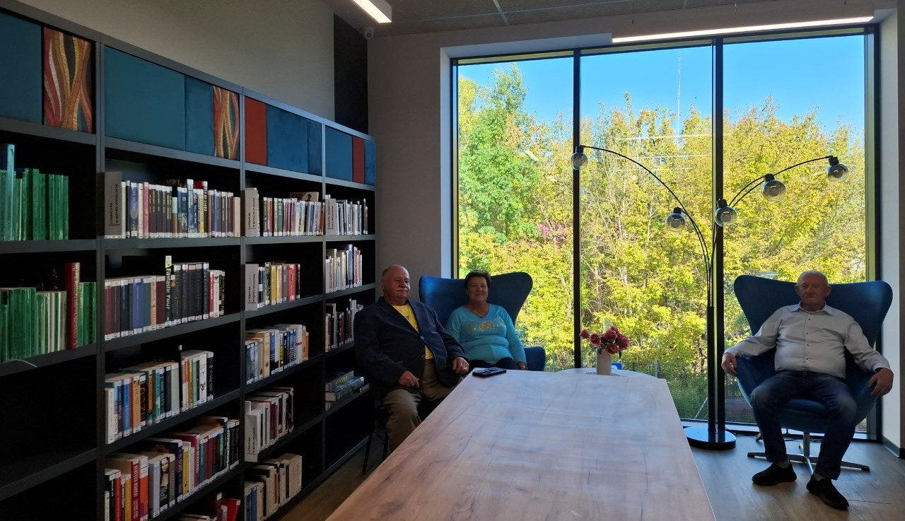 20260928_143324 Trzy osoby siedzące na krzesłach przed dużym oknem, w jasnym pomieszczeniu biblioteki.