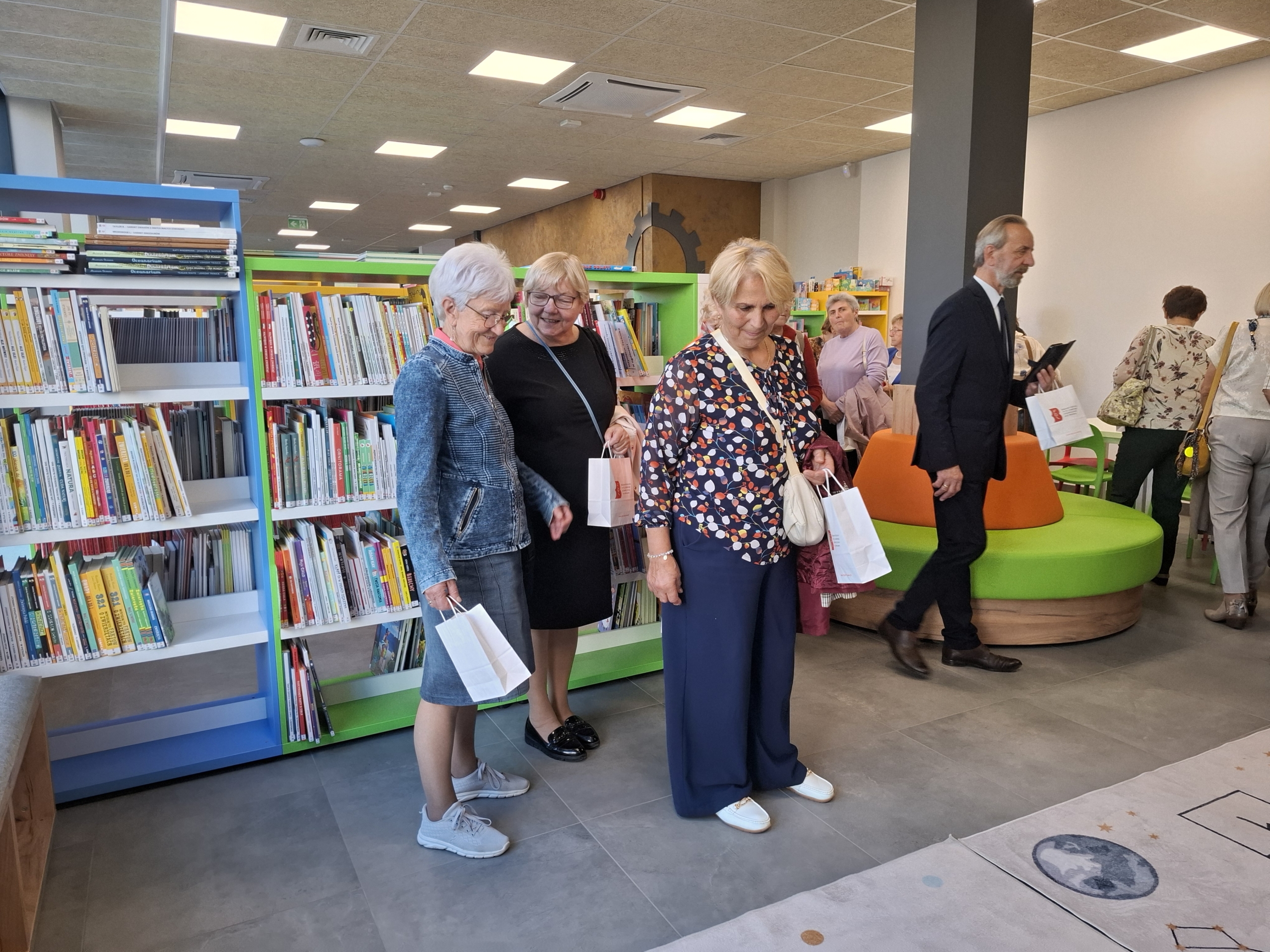 20260928_144413 Grupa ludzi stojących w bibliotece, przeglądających książki i rozmawiających w cichym otoczeniu.